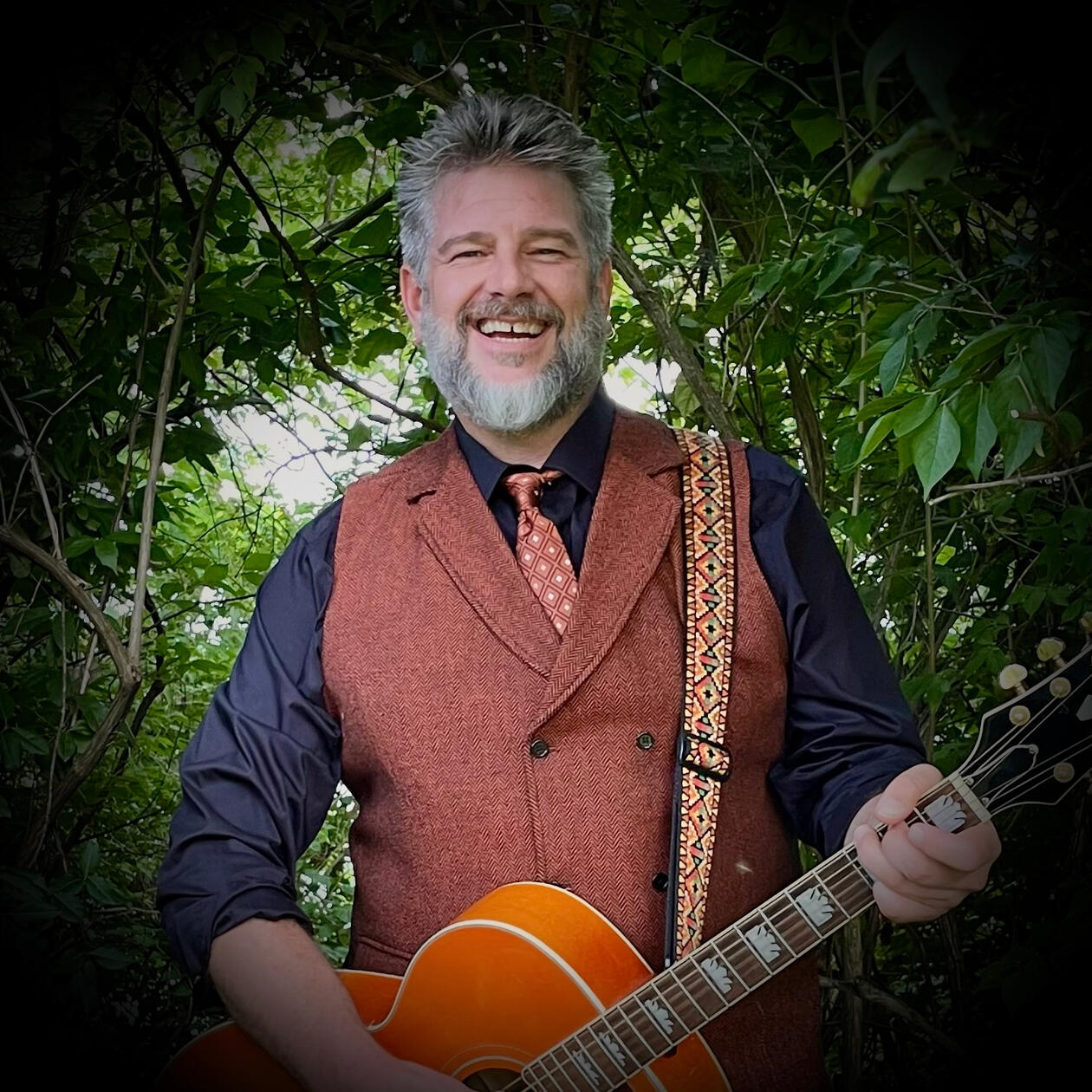 Mr. Singer – Beloved Kids' Performer in Nashville Smiling Mr. Singer with guitar in orange vest and black shirt, standing in greenery